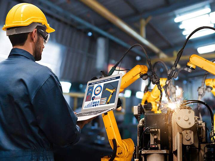 Worker in blue coveralls and yellow hardhat monitoring a laptop of connected to industrial robot arms in process