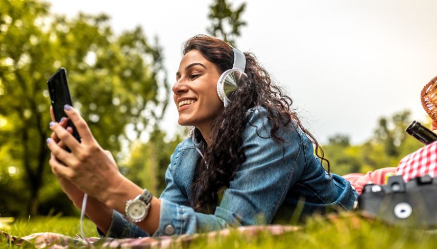 Woman lying on grass outdoors, smiling at her phone while wearing headphones.